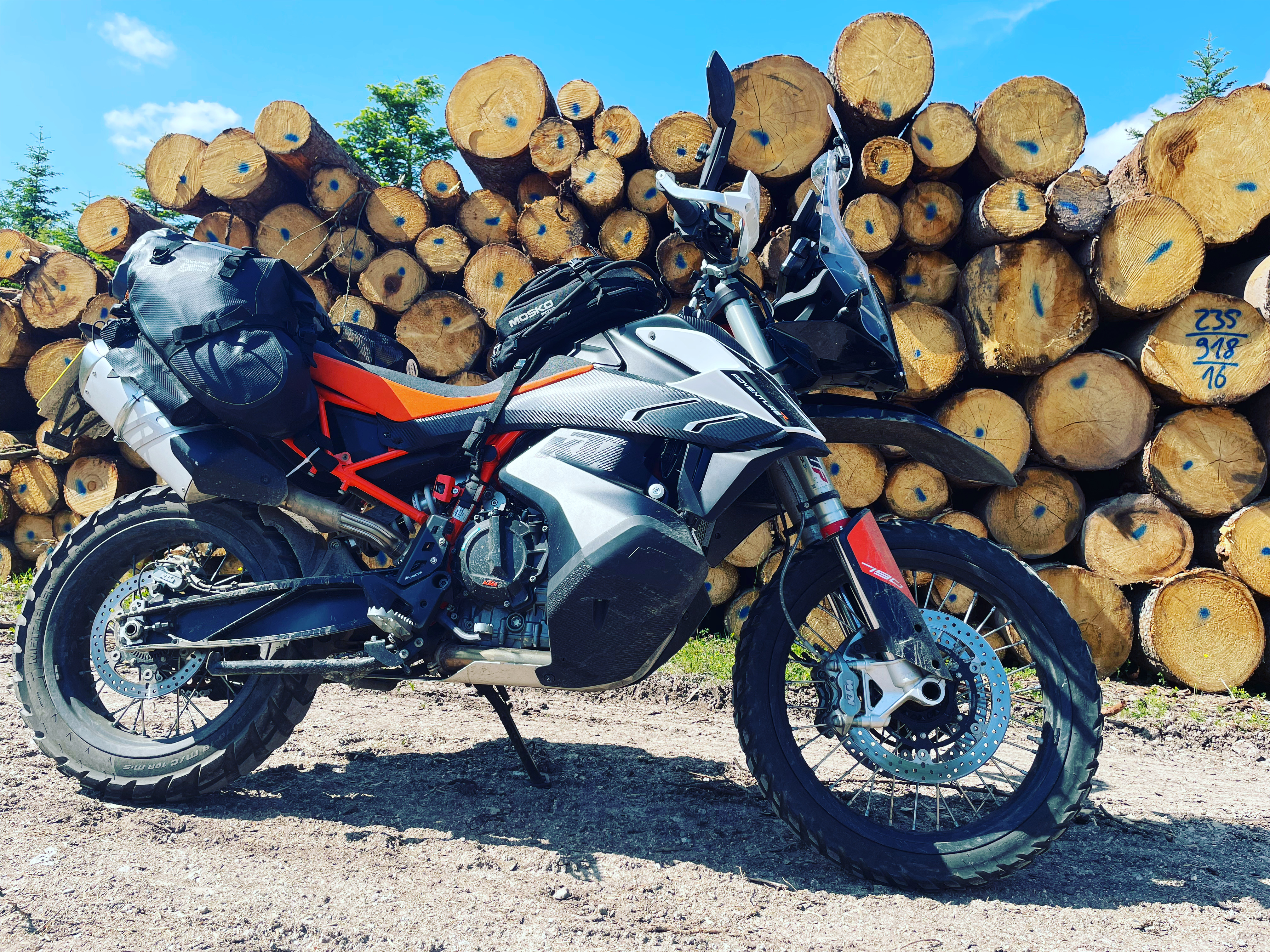 A KTM 790 Adventure R parked in front of a stack of logs under a blue sky.